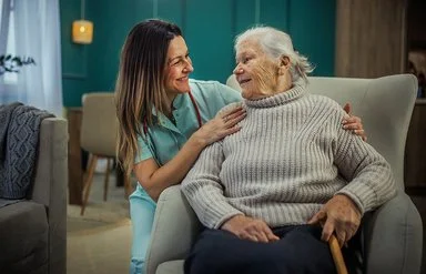 Eine alte Frau sitzt in einem Sessel. Neben ihr kniet eine Pflegekraft und legt ihre Hände auf die Schultern der Frau. Sie sehen sich in die Augen und lächeln.