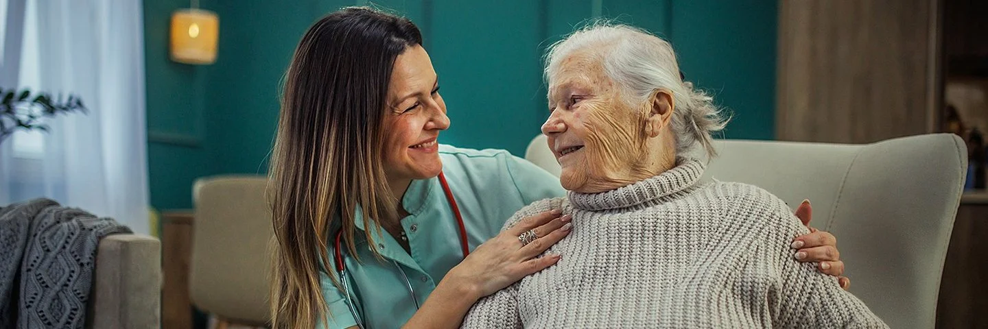 Eine alte Frau sitzt in einem Sessel. Neben ihr kniet eine Pflegekraft und legt ihre Hände auf die Schultern der Frau. Sie sehen sich in die Augen und lächeln.