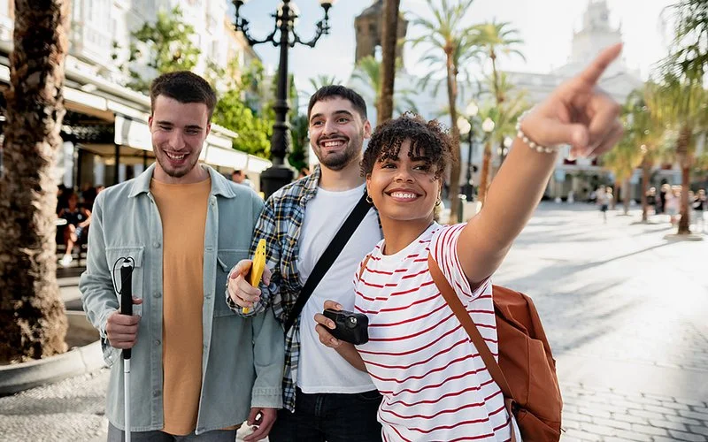 Eine Gruppe junger Menschen steht in einer Innenstadt. Eine junge Frau zeigt auf etwas, ein Mann hält einen Langstock in der Hand.