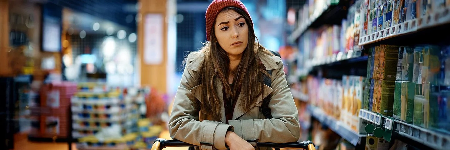 Eine Frau schiebt einen Einkaufswagen und schaut kritisch auf die Supermarktregale.