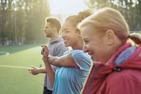 Drei Personen im Alter von etwa 25 bis 65 Jahren machen in Sportbekleidung Dehnübungen auf einem Sportplatz in der Morgensonne.