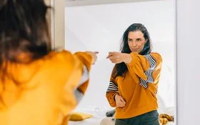 Eine Frau mit dunklen langen Haaren und einem auffälligen orange-gelben Oberteil zeigt selbstbewusst auf ihr Spiegelbild und blickt dabei sehr glücklich und zufrieden.