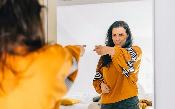 Eine Frau mit dunklen langen Haaren und einem auffälligen orange-gelben Oberteil zeigt selbstbewusst auf ihr Spiegelbild und blickt dabei sehr glücklich und zufrieden.