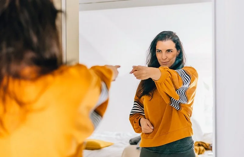 Eine Frau mit dunklen langen Haaren und einem auffälligen orange-gelben Oberteil zeigt selbstbewusst auf ihr Spiegelbild und blickt dabei sehr glücklich und zufrieden.