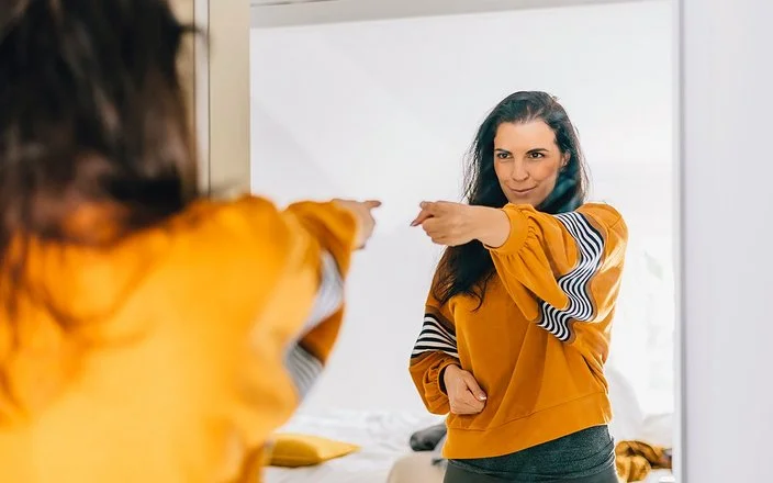 Eine Frau mit dunklen langen Haaren und einem auffälligen orange-gelben Oberteil zeigt selbstbewusst auf ihr Spiegelbild und blickt dabei sehr glücklich und zufrieden.