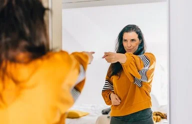 Eine Frau mit dunklen langen Haaren und einem auffälligen orange-gelben Oberteil zeigt selbstbewusst auf ihr Spiegelbild und blickt dabei sehr glücklich und zufrieden.