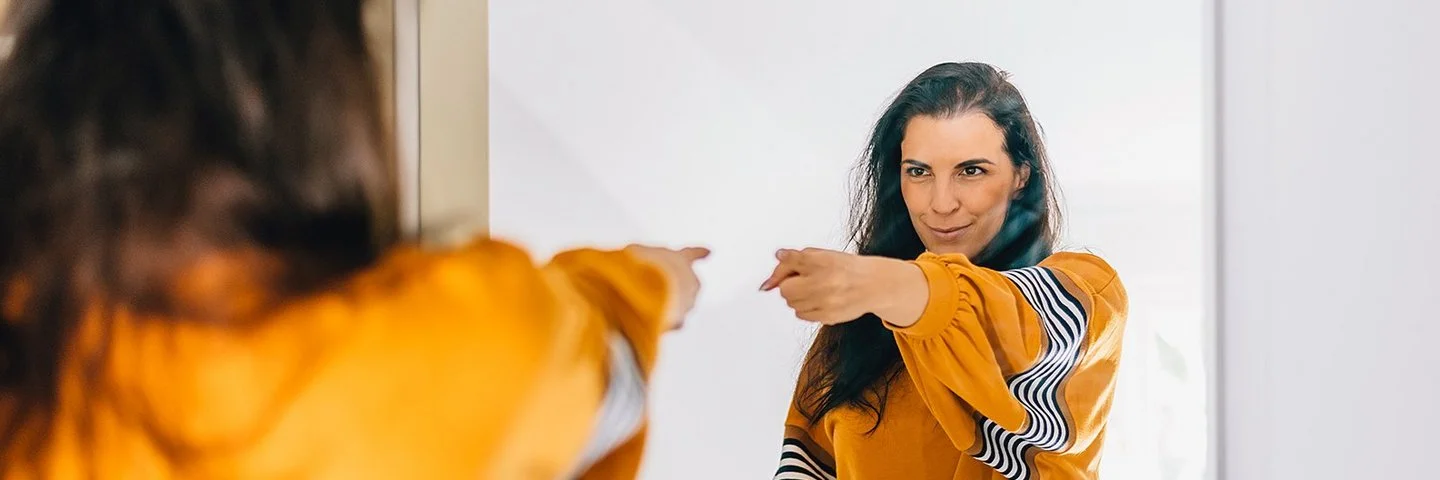 Eine Frau mit dunklen langen Haaren und einem auffälligen orange-gelben Oberteil zeigt selbstbewusst auf ihr Spiegelbild und blickt dabei sehr glücklich und zufrieden.