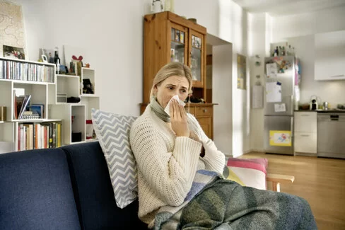 Eine Frau sitzt am Sofa und putzt ihre Nase. Hinter ihren Beschwerden könnte eine Allergie gegen Hausstaubmilben stecken. 