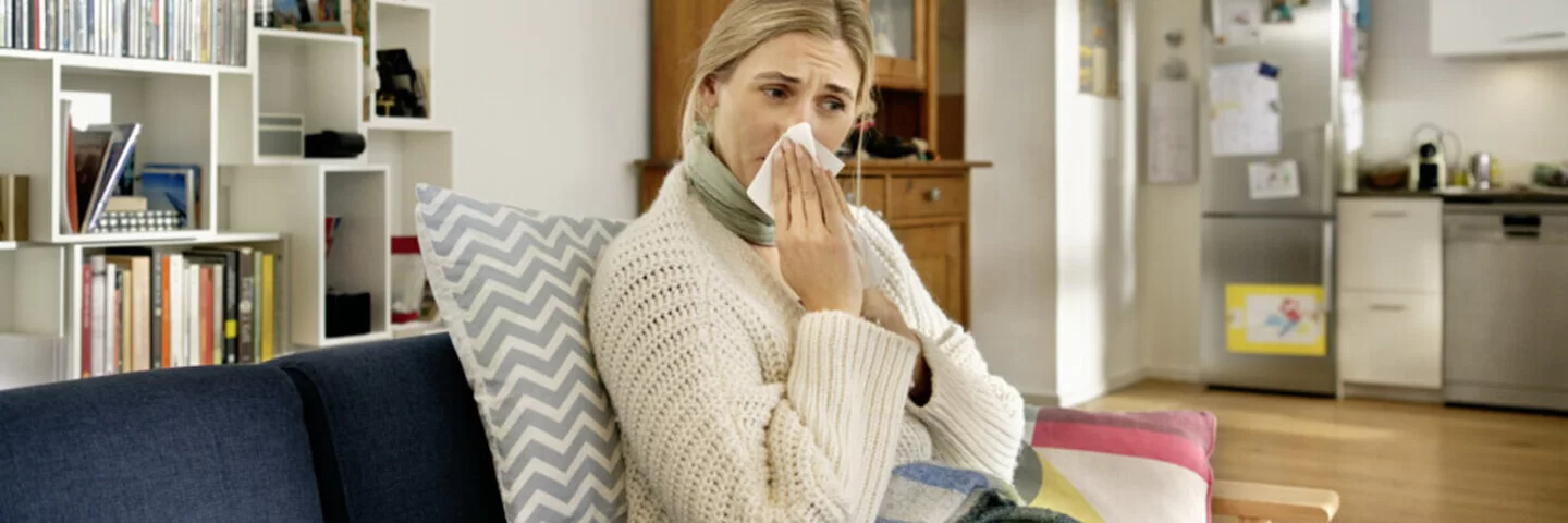 Eine Frau sitzt am Sofa und putzt ihre Nase. Hinter ihren Beschwerden könnte eine Allergie gegen Hausstaubmilben stecken. 