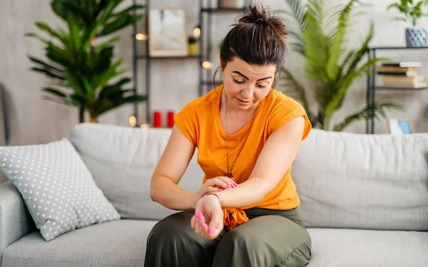Eine junge Frau in einem Wohnzimmer. Sie sitzt auf einem Sofa und betrachtet die Innenseite ihres linken Unterarms. Dort ist ein schwach roter Strich auf der Haut zu sehen, der sich vom Handgelenk bis zum Ellenbogen zieht. Ihr Blick ist leicht besorgt.