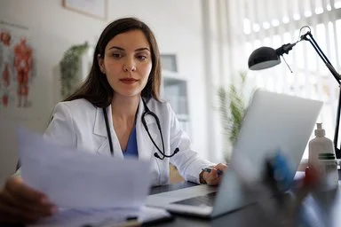 Eine Ärztin im weißen Kittel mit Stethoskop sitzt an einem Schreibtisch mit Laptop und betrachtet konzentriert ein Dokument.