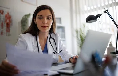Eine Ärztin im weißen Kittel mit Stethoskop sitzt an einem Schreibtisch mit Laptop und betrachtet konzentriert ein Dokument.