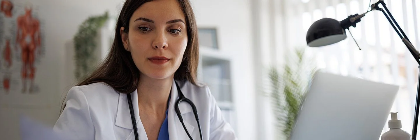 Eine Ärztin im weißen Kittel mit Stethoskop sitzt an einem Schreibtisch mit Laptop und betrachtet konzentriert ein Dokument.