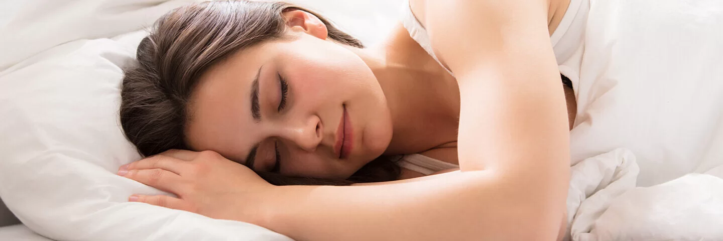 Gesunder Schlaf: Eine junge Frau liegt auf der Seite in einem weiß bezogenen Bett und schläft entspannt. 