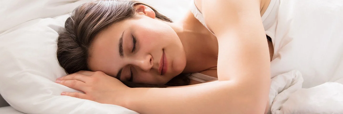 Gesunder Schlaf: Eine junge Frau liegt auf der Seite in einem weiß bezogenen Bett und schläft entspannt.
