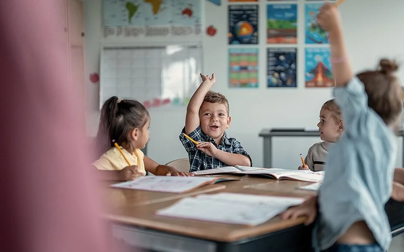 In einem Klassenzimmer sitzen Grundschulkinder an ihren Schultischen. In der Bildmitte hebt ein Junge lächelnd den Arm. Die beiden Mädchen, die links und rechts von ihm sitzen, blicken ihn an. Rechts steht ein weiteres Mädchen, das sich ebenfalls meldet.
