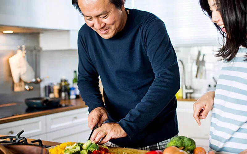 Ein Mann steht in der Küche und schneidet Gemüse, eine Frau sieht ihm dabei zu und lacht.