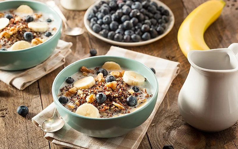 Eine Frühstücksbowl mit Quinoa, Nüssen, Blaubeeren und Bananenscheiben auf einem Holztisch. Daneben steht eine kleine Milchkanne, im Hintergrund sind eine Banane, Blaubeeren und eine weitere Bowl zu sehen.
