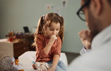 Ein kleines Mädchen sitzt in einem Zimmer und hustet in die rechte Hand. In der linken Hand hält das Mädchen ein Kuscheltier. Der behandelnde Arzt sitzt vor ihr und ist nur verschwommen zu erkennen. 