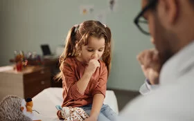 Ein kleines Mädchen sitzt in einem Zimmer und hustet in die rechte Hand. In der linken Hand hält das Mädchen ein Kuscheltier. Der behandelnde Arzt sitzt vor ihr und ist nur verschwommen zu erkennen. 