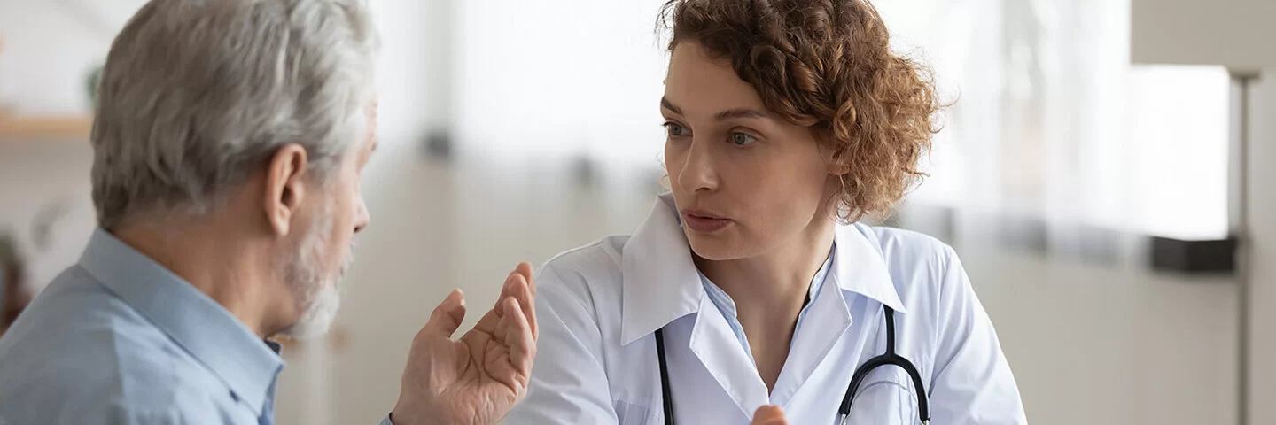 Eine Ärztin spricht mit einem älteren männlichen Patienten. Der Patient sitzt entspannt und hört aufmerksam zu, während die Ärztin ihm etwas erklärt.