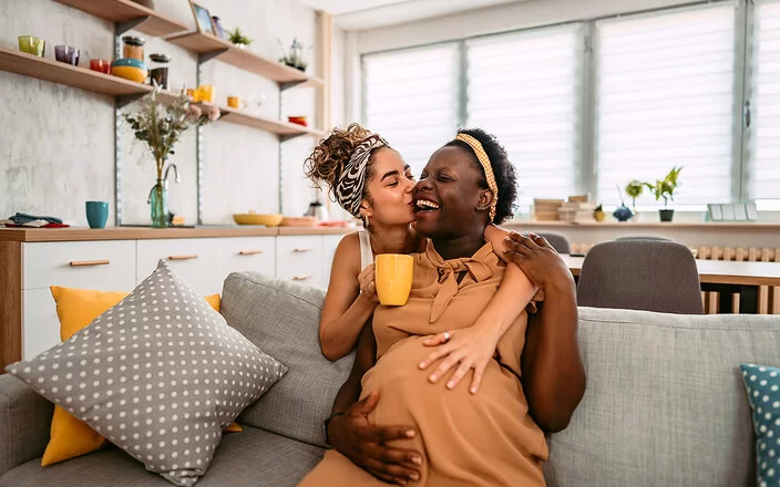 Eine junge Frau sitzt lachend auf dem Sofa und wird von ihrer Partnerin von hinten umarmt und auf die Wange geküsst.