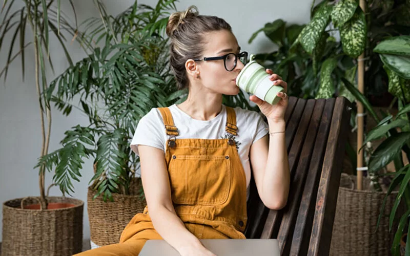 Eine junge Frau im Overall sitzt auf einem Stuhl und trinkt aus einem wiederverwendbaren Kaffeebecher.