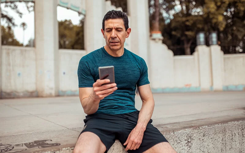 Ein sportlicher Mann mittleren Alters sitzt auf dem Boden und schaut auf sein Smartphone.