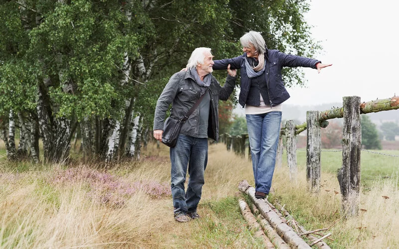 Älteres Paar geht ein Feld entlang, sie balanciert auf Holzstämmen, er stützt sie dabei.