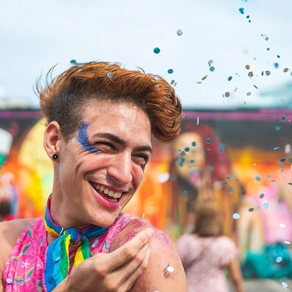 Porträt eines lachenden queeren Mannes auf einem Festival, der blauen Glitzer neben seinem rechten Auge trägt und Konfetti in die Luft wirft.