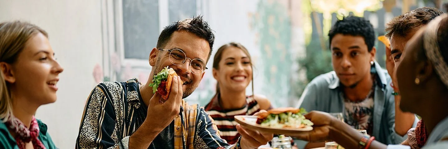 Sechs Frauen und Männer sitzen zusammen am Tisch und essen Tacos, die Stimmung ist fröhlich.