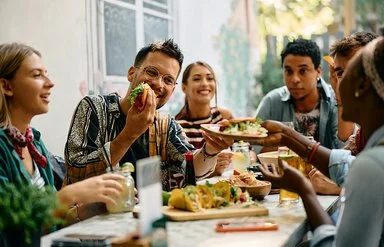 Sechs Frauen und Männer sitzen zusammen am Tisch und essen Tacos, die Stimmung ist fröhlich.