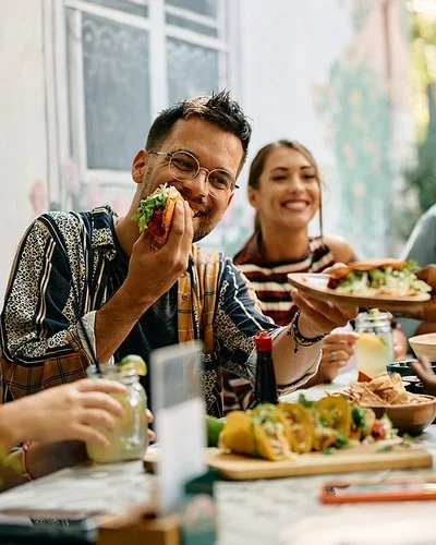 Sechs Frauen und Männer sitzen zusammen am Tisch und essen Tacos, die Stimmung ist fröhlich.