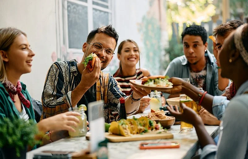Sechs Frauen und Männer sitzen zusammen am Tisch und essen Tacos, die Stimmung ist fröhlich.