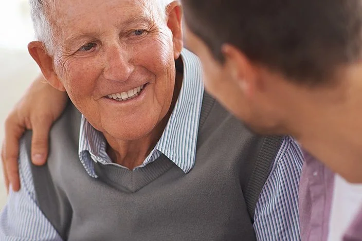 Ein Mann legt den Arm um die Schulter seines alten Vaters. Sie sitzen zusammen auf einer hellen Couch und reden miteinander.