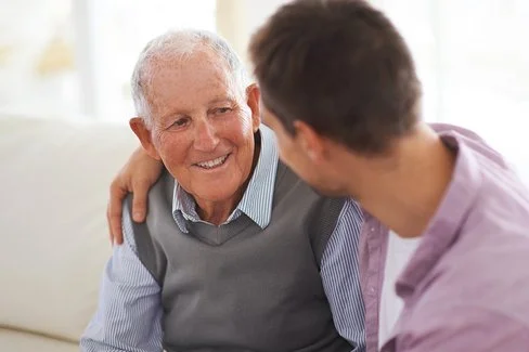 Ein Mann legt den Arm um die Schulter seines alten Vaters. Sie sitzen zusammen auf einer hellen Couch und reden miteinander.