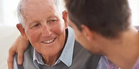 Ein Mann legt den Arm um die Schulter seines alten Vaters. Sie sitzen zusammen auf einer hellen Couch und reden miteinander.