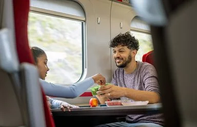 Ein junger Mann und ein Mädchen sitzen sich im Zug gegenüber. Sie greift zu einem frischen Snack in einer Aufbewahrungsdose, die er ihr hinhält.