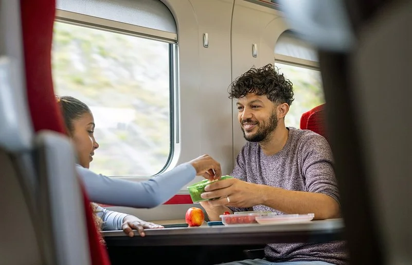 Ein junger Mann und ein Mädchen sitzen sich im Zug gegenüber. Sie greift zu einem frischen Snack in einer Aufbewahrungsdose, die er ihr hinhält.