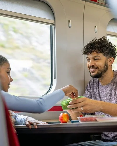 Ein junger Mann und ein Mädchen sitzen sich im Zug gegenüber. Sie greift zu einem frischen Snack in einer Aufbewahrungsdose, die er ihr hinhält.