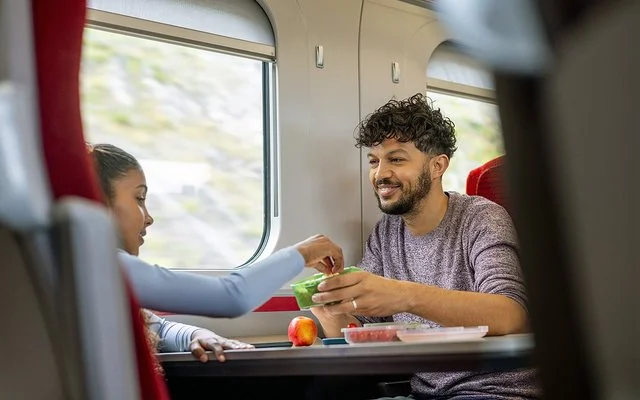 Ein junger Mann und ein Mädchen sitzen sich im Zug gegenüber. Sie greift zu einem frischen Snack in einer Aufbewahrungsdose, die er ihr hinhält.