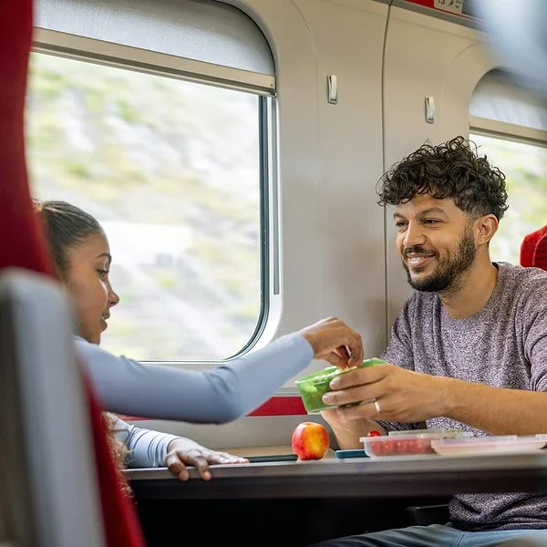 Ein junger Mann und ein Mädchen sitzen sich im Zug gegenüber. Sie greift zu einem frischen Snack in einer Aufbewahrungsdose, die er ihr hinhält.