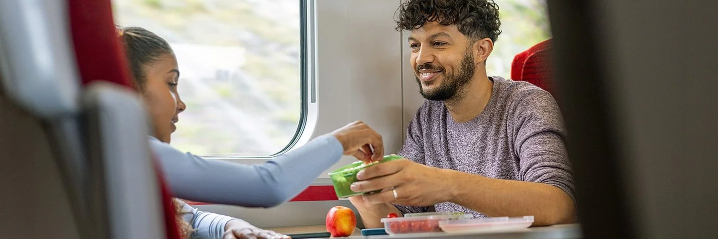 Ein junger Mann und ein Mädchen sitzen sich im Zug gegenüber. Sie greift zu einem frischen Snack in einer Aufbewahrungsdose, die er ihr hinhält.