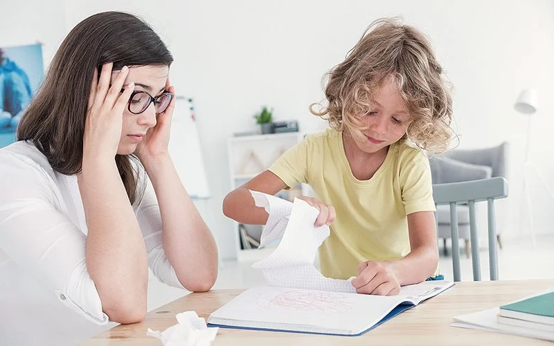 Eine Mutter sitzt mit aufgestützten Armen am Tisch und fasst sich mit beiden Händen an die Schläfen, während ihr Kind Seiten aus einem Heft reißt.