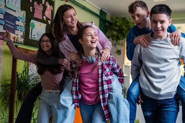 Eine Gruppe von fünf Jugendlichen in einem Flur, zwei tragen jeweils eine andere Person auf dem Rücken, alle lachen ausgelassen.
