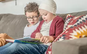 Ein gesundes Kind sitzt mit seinem erkrankten Geschwisterkind auf der Couch und liest ein Buch.