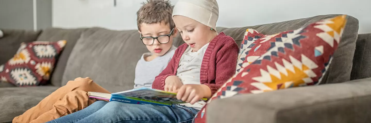 Ein gesundes Kind sitzt mit seinem erkrankten Geschwisterkind auf der Couch und liest ein Buch.