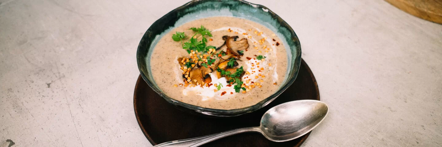 Champignoncremesuppe mit knusprigem Buchweizentopping nach einem Rezept von Felicitas Then, angerichtet auf einem schwarzen Teller, garniert mit Petersilie und Sauerrahm.