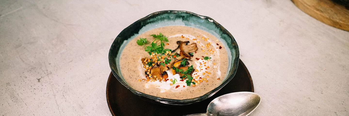 Champignoncremesuppe mit knusprigem Buchweizentopping nach einem Rezept von Felicitas Then, angerichtet auf einem schwarzen Teller, garniert mit Petersilie und Sauerrahm.