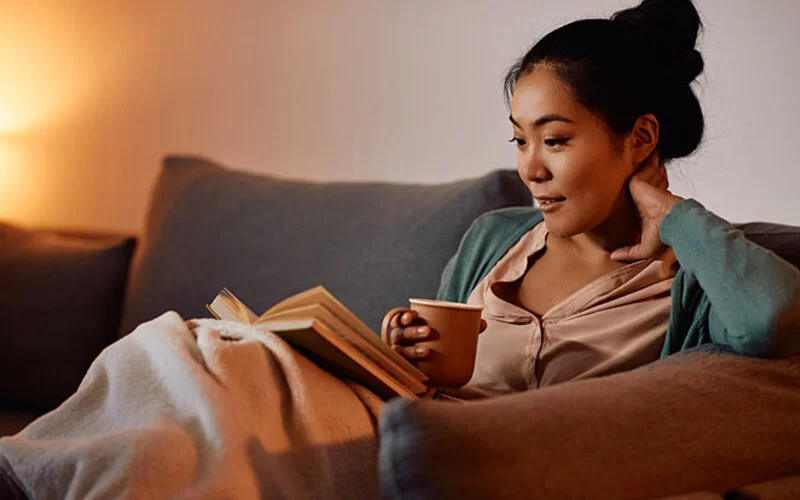 Eine Frau liest vor dem Einschlafen ein Buch und trinkt eine Tasse Tee – das hilft ihr, besser einzuschlafen.